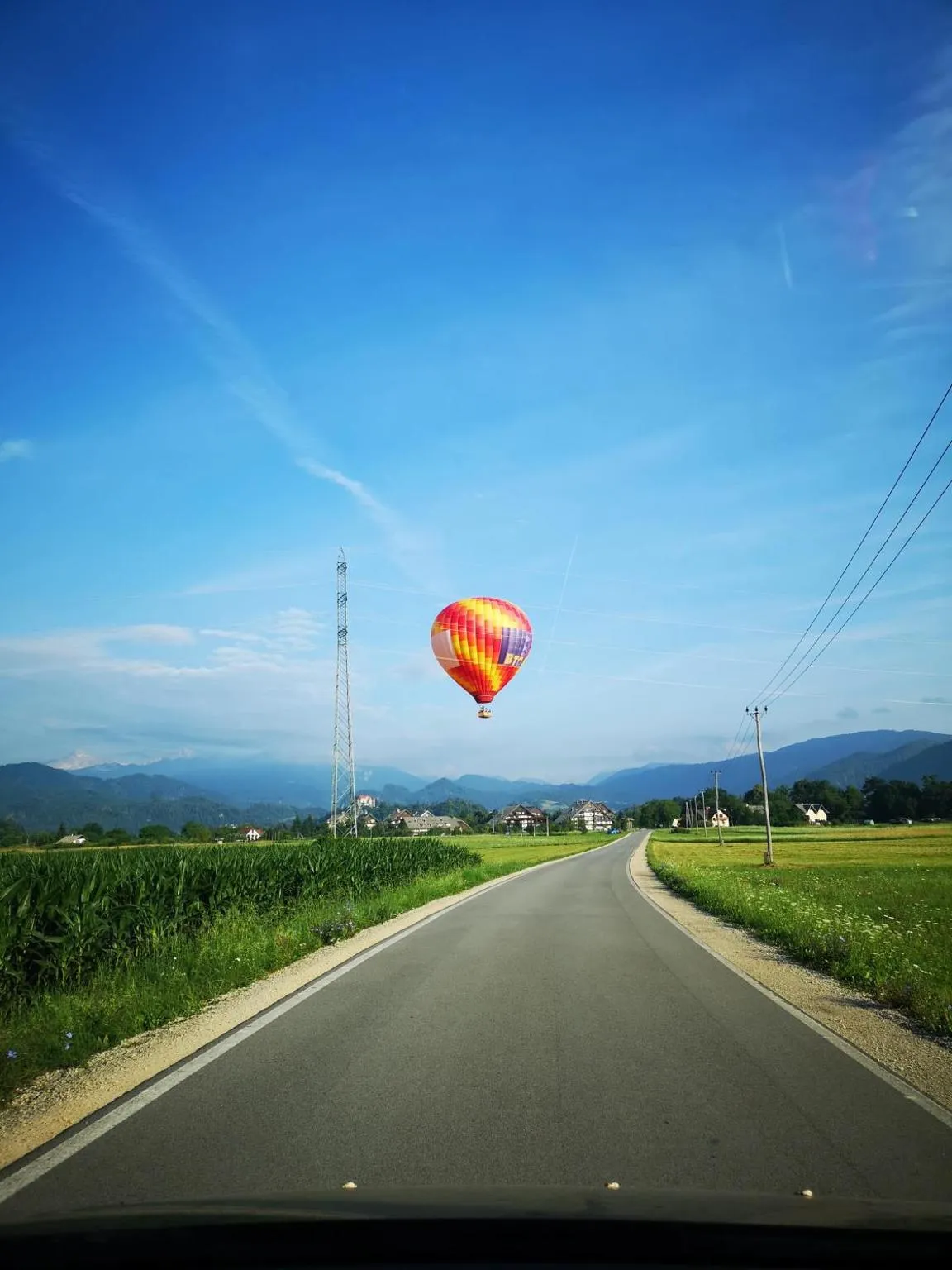 Cesta med koruzo in travniki proti vasi, nad njo balon, v ozadju gore